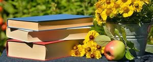 Pile of three books on table with apple and metal pot of yellow flowers