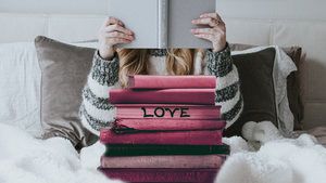 Girl reading in bed, grey book hides her face as she reads. In foreground is a pile of red books and on one spine is the word Love.
