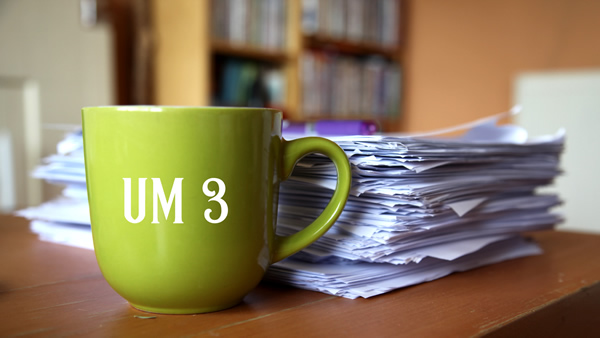 Chartreuse green mug with text UM3 in white on the side, next to a tall pile of A4 sheets of paper