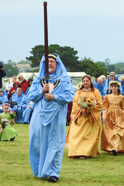 Bard in blue robes carrying an upright sheathed sword with The Lady of Cornwall dressed in pale orange carrying a bouquet of wheat with her attendants. Circle of blue robed bards in the background.