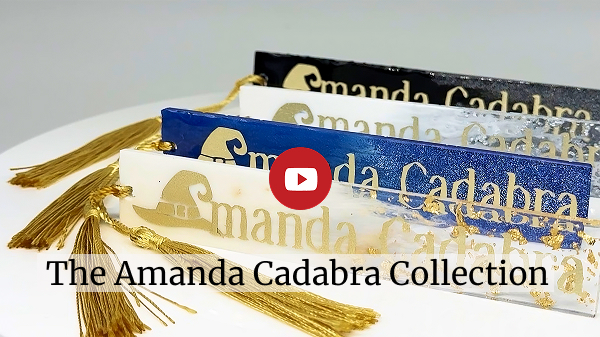 4 resin craft books mark: one gold foil in white resin, silver foil in white, laster royal blue and black with holographic glitter, each with a gold tassel arranged in a row o their edges, each has the words Amanda Cadabra in gold inset. Click to go to video of the Amanda Cadabra Collection