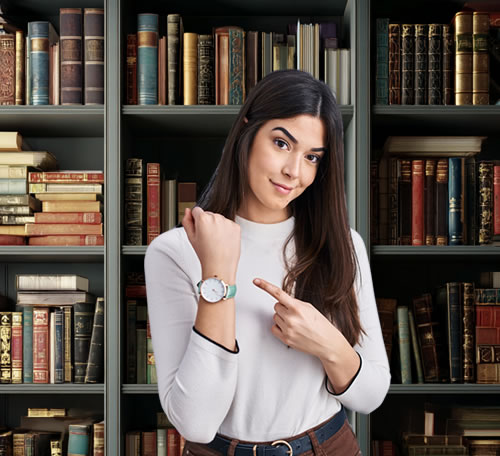 Woman with long dark hair wearing white long sleeve top pointing to wrist watch with smiling questioning look at camera