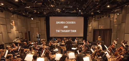 Film studio orchestra with screen in the background with text: Amanda Cadabra and The Twilight Toxin