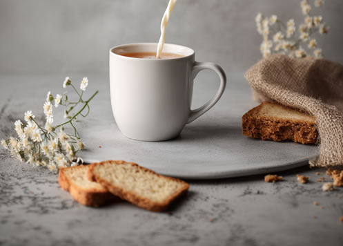 milk streaming into cup of tea with dried flowers, cake, toast and unbleached linen around it