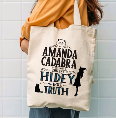 Tote bag on the shoulder of a woman in an organge top. Cream background tote with Text: Amanda Cadabra and The Hidey-Hole Truth with silouette of Amanda in witch hat on the right looking up and silhouette of Tempest her cat on the bottom right